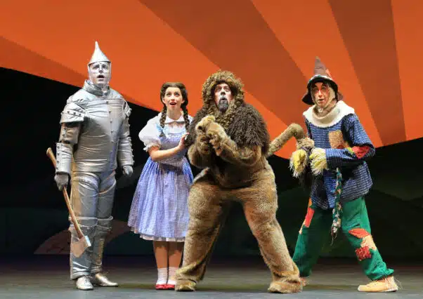 Actors in costume performing as the Tin Man, Dorothy, the Cowardly Lion, and the Scarecrow from 'The Wizard of Oz' on stage, with a colorful orange and black backdrop.