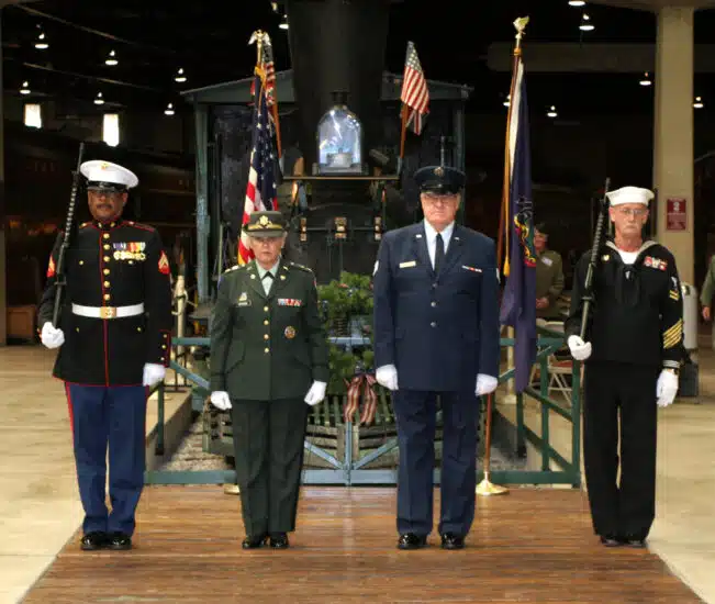 Four U.S. military service members stand at attention in uniform during a ceremony indoors. Each represents a different branch of the Armed Forces — Marine Corps, Army, Air Force, and Navy — positioned in front of an American flag and a historic train engine decorated with a wreath and flags.