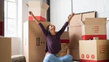 Happy young woman sitting in new apartment and raising arms in joy after moving in.