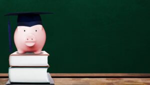 Saving for education with pink piggy bank wearing graduate hat on books in school classroom with copy space on chalkboard. Concept of education investment, tuition cost, college loan, etc.