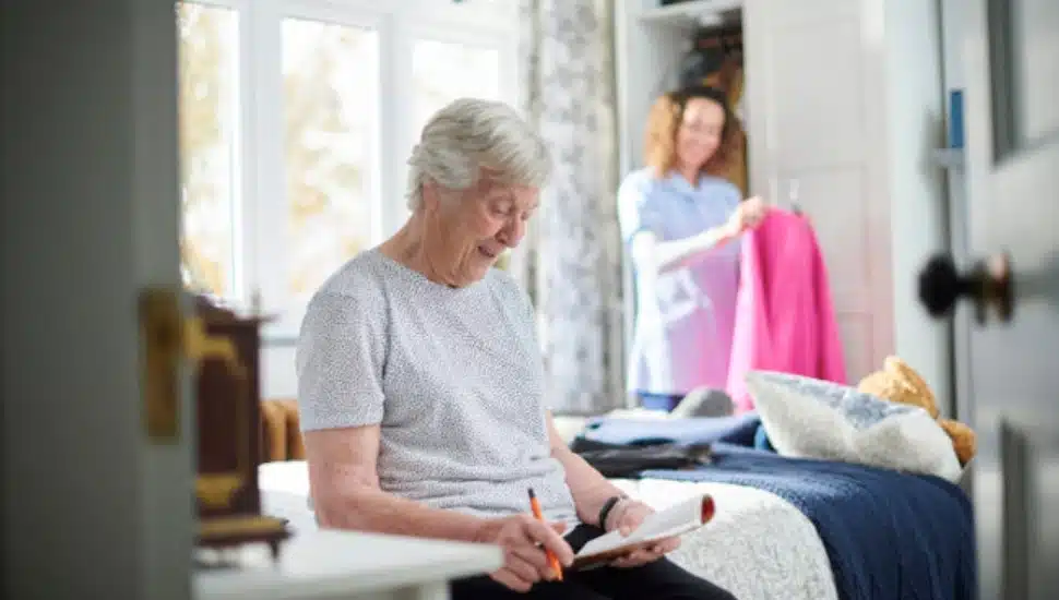 An elderly woman writing on a notepad while sitting on the bed with a nurse or aide hanging up clothes in the background.