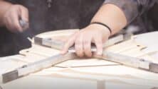 front-facing view of craftsperson carving wooden beams in cross-shaped pattern behind front piece of guitar