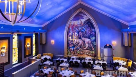 Interior of Oldestone Steakhouse, tables and chairs in dining area. Walls adorned with stained glass windows and mural