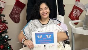 Dana Nieves of St. Mary Medical Center in hospital gown with newborn baby girl holding certificate in front of Christmas tree and stockings
