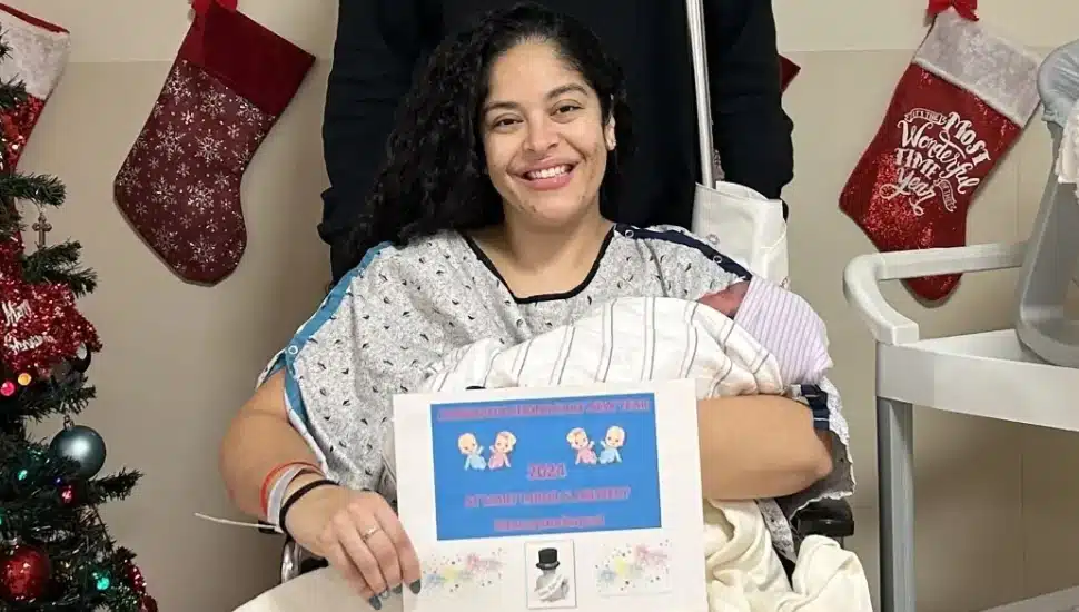 Dana Nieves of St. Mary Medical Center in hospital gown with newborn baby girl holding certificate in front of Christmas tree and stockings