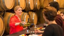 woman pouring wine in guests' glasses at winery in front of barrels