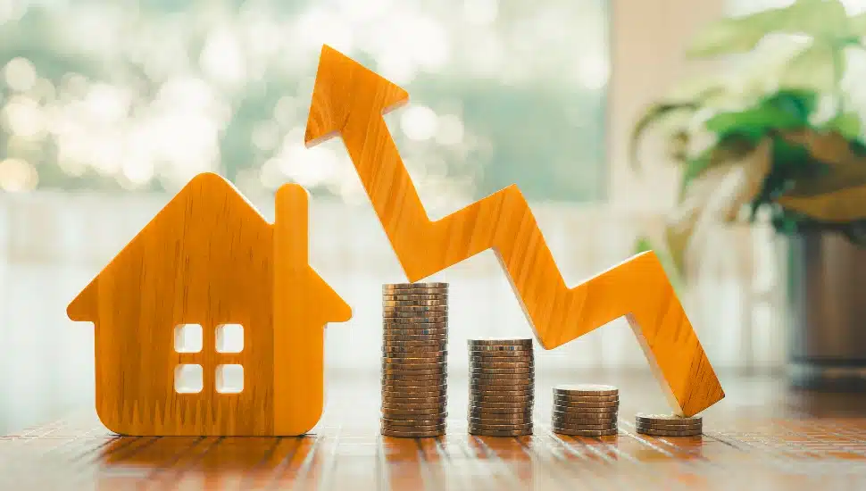 image of wooden house figurine on table next to four stacks of coins progressively increasing and a wooden upwards left zig zag arrow