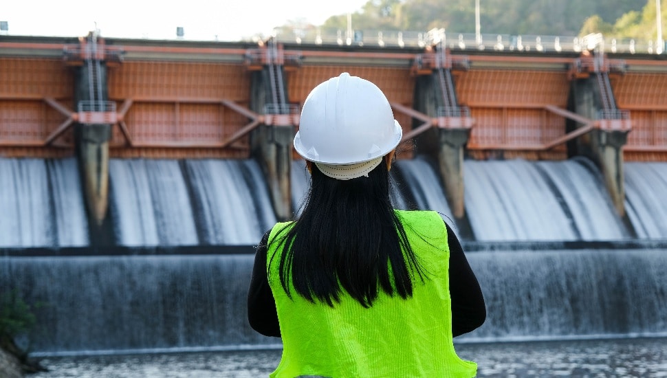 Rear view of female engineer in green vest and helmet standing outside against background of dam with hydroelectric power plant and irrigation.