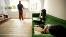 Father stands, looking out the window with two young daughters sitting on the couch behind him.