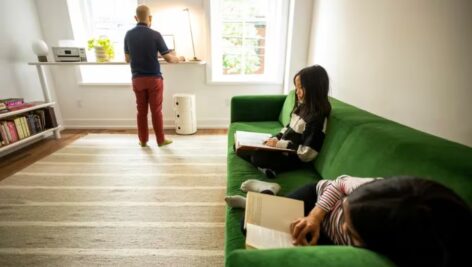 Father stands, looking out the window with two young daughters sitting on the couch behind him.