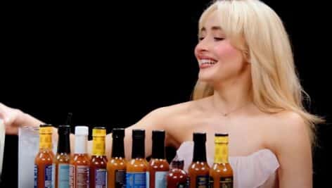 Sabrina Carpenter sitting at table with bottles of hot sauce