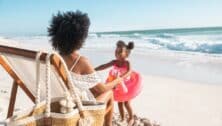 Black cute little girl inside pink float standing on beach with mother while playing together. Rear view of young african american mother sitting on deck chair holding hands of her daughter standing with flamingo inflatable for swim at beach. Young woman with child having fun together at sea.