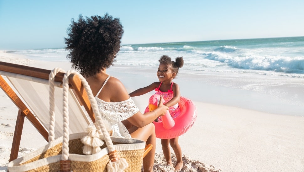 Black cute little girl inside pink float standing on beach with mother while playing together. Rear view of young african american mother sitting on deck chair holding hands of her daughter standing with flamingo inflatable for swim at beach. Young woman with child having fun together at sea.