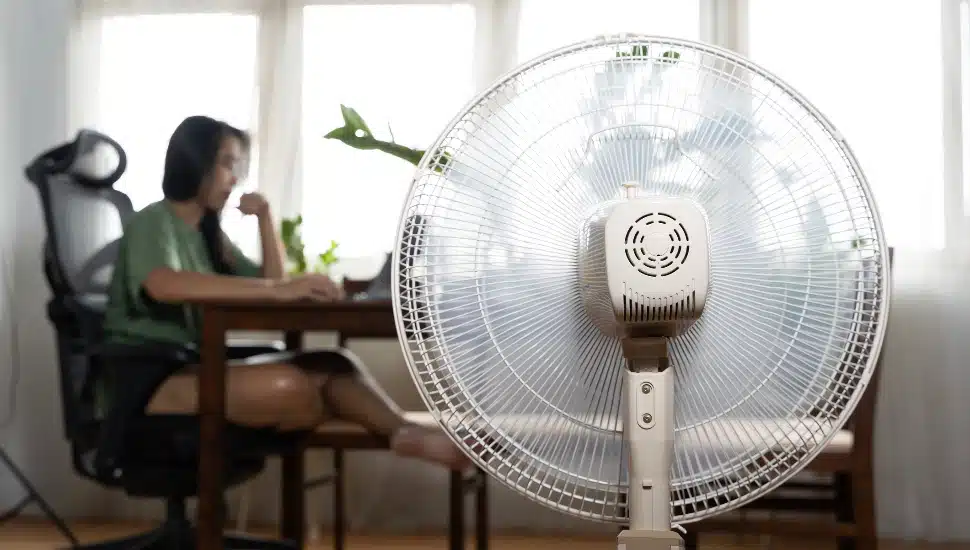 Sunny summer in front of the working fan, suffering from the summer heat. Unhappy Asian woman sitting in front of fan at work suffering from heat in modern house, enjoying cooling wind.