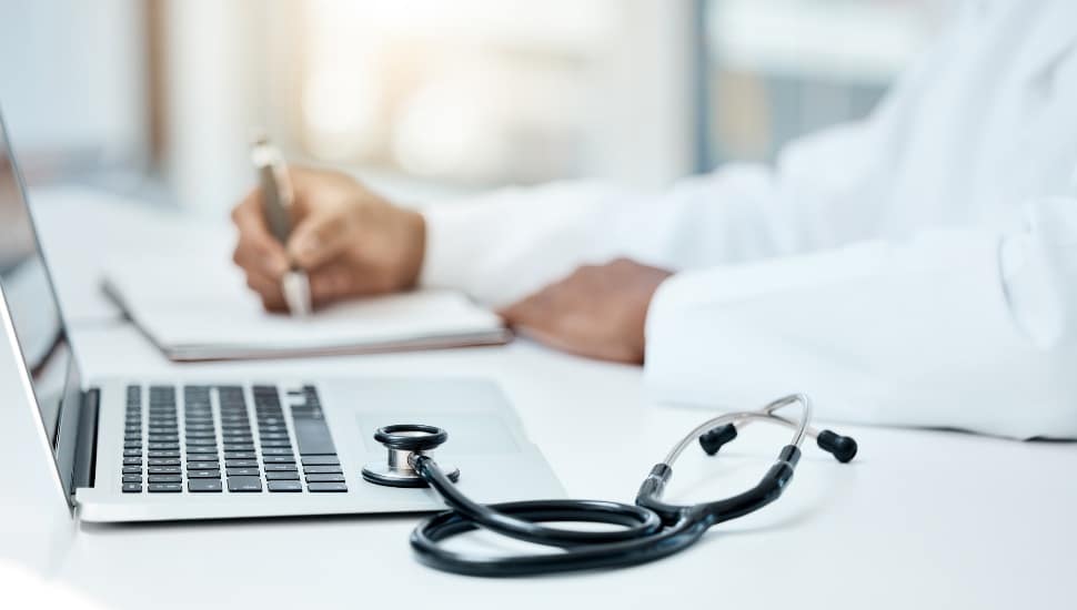 laoptop on desk with doctor's forearms resting on desk and stethoscope