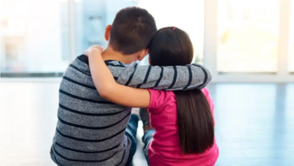 siblings putting arms around eachother, sitting on floor looking out window