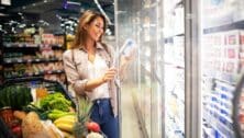 Woman with shopping cart opening supermarket freezer choosing what to buy.