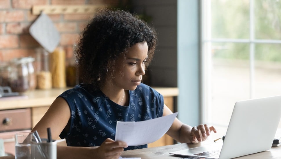 Serious millennial African American woman calculating taxes, budget, costs, checking paper invoice, receipt. Homeowner, tenant using laptop for paying bill, rent, mortgage, insurance fees.