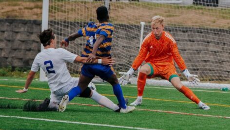 Soccer action shot: a player in a white uniform slides to tackle the ball from an opponent in blue and yellow stripes, as an alert goalkeeper in an orange jersey prepares to block in front of the goal. The intensity of the play highlights the competitive nature of the game.