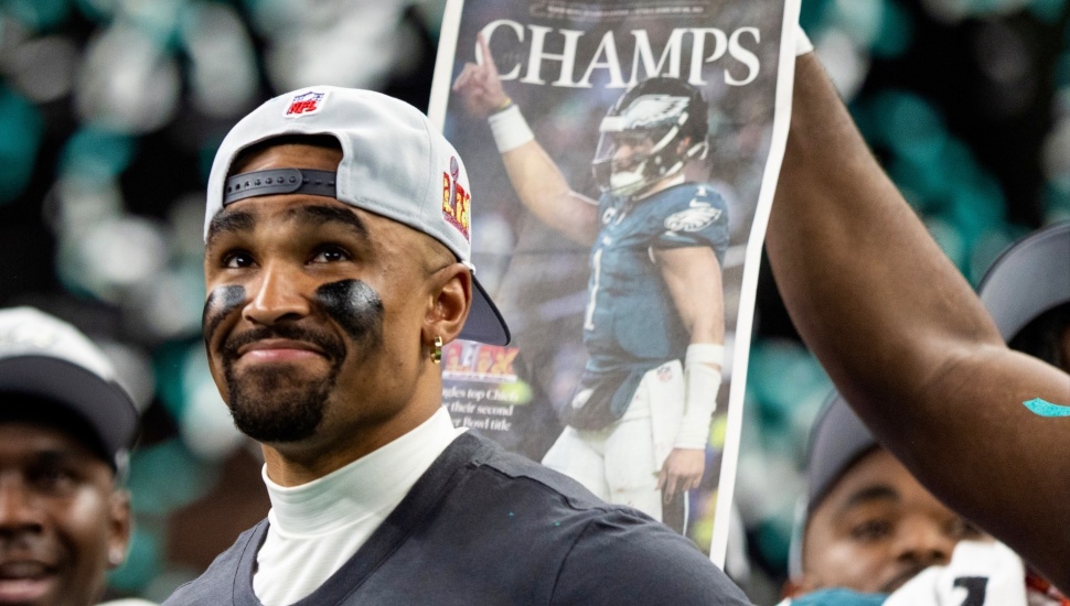 Jalen Hurts smiling with Eagles Super Bowl hat and shirt on. A newspaper with "Champs"