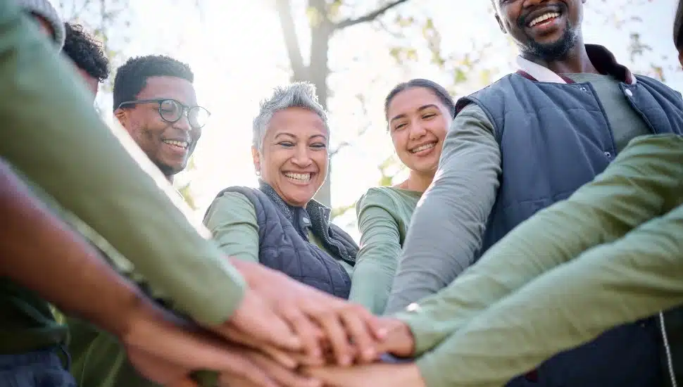A diverse group of people smiling and joining hands in a circle outdoors, symbolizing teamwork, community, and unity.