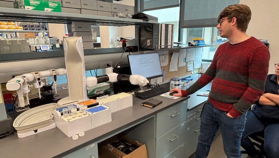 A lab technician stands next to a high-tech robotic arm system used for PFAS testing at Aqua Pennsylvania’s laboratory. The robot is set up on a lab bench alongside test tubes and a computer interface.