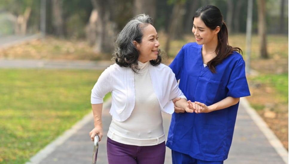 A nurse or health care worker helps an older woman with MS during a walk on a path.