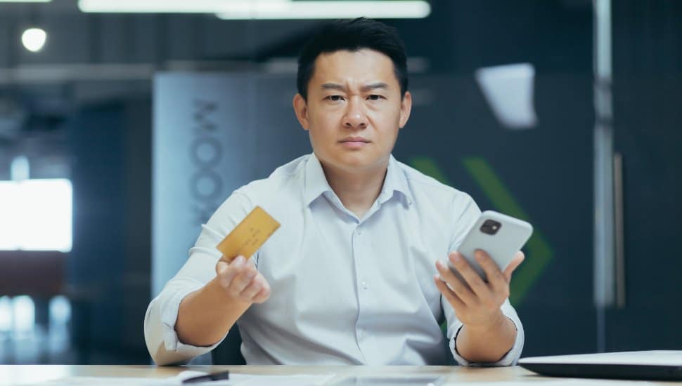 A frustrated man holding credit card and phone at a table, disgruntled with his bank.