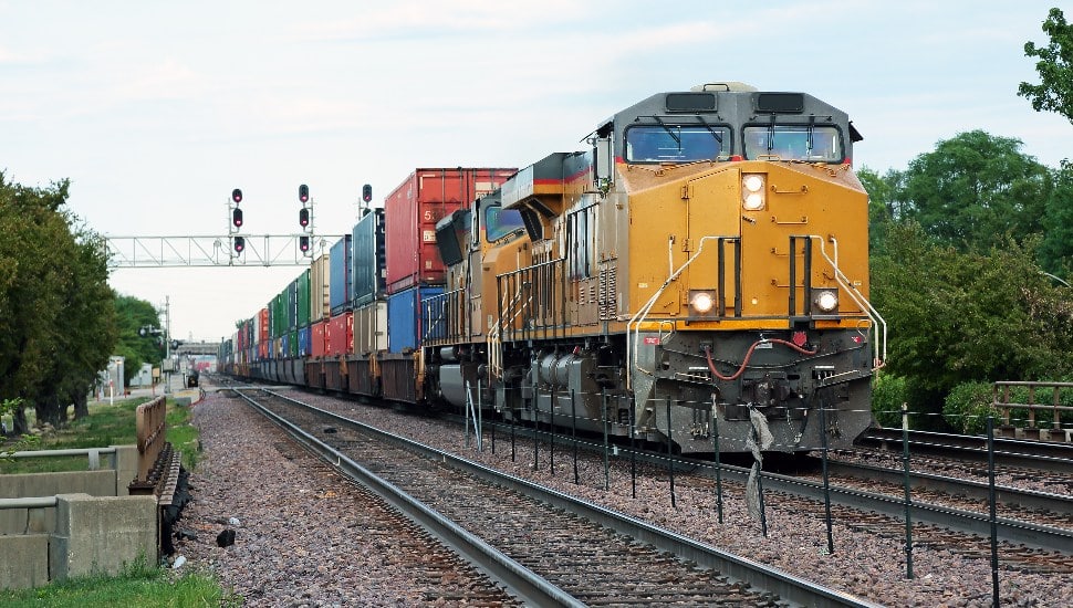 Two yellow locomotives and double stack freight train passing through town.
