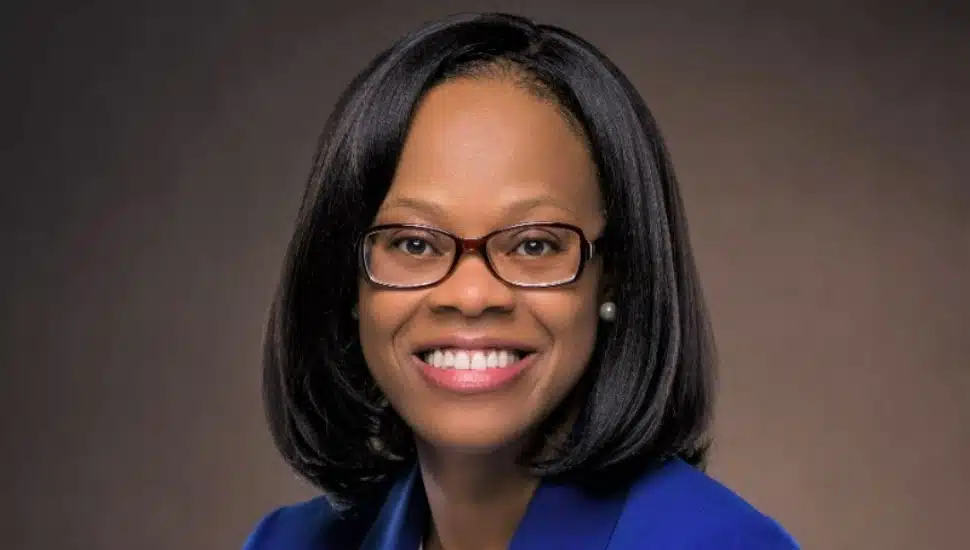 Professional headshot of a smiling woman with straight shoulder-length hair, wearing glasses, pearl earrings, and a royal blue blazer, posed against a neutral brown background.