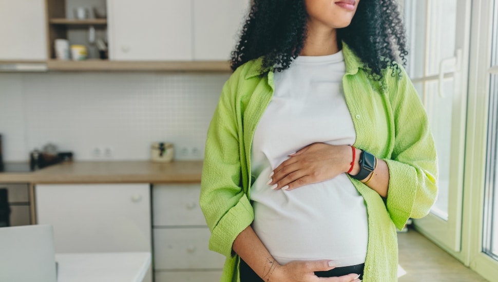No face picture of pregnant African American woman with long curly hair looking through window standing in kitchen, rubbing her pregnant belly.
