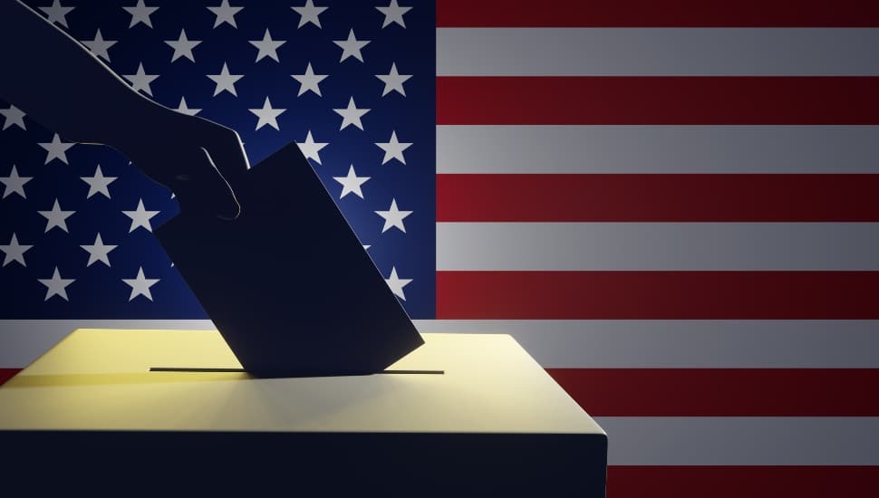 Silhouette of a hand putting vote in ballot box with an American flag in the background.