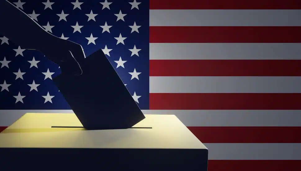 Silhouette of a hand putting vote in ballot box with an American flag in the background.