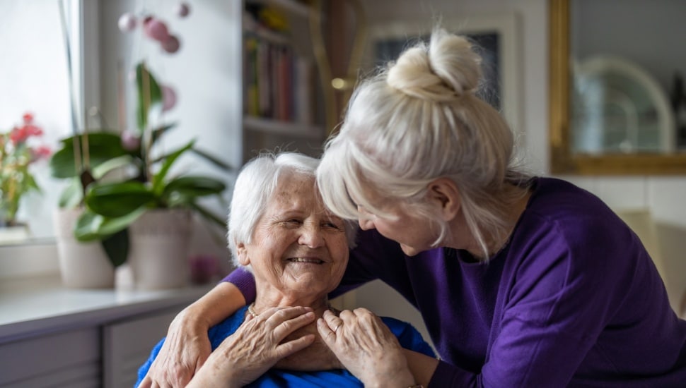 A woman provides comfort to an elderly woman sitting in a chair.