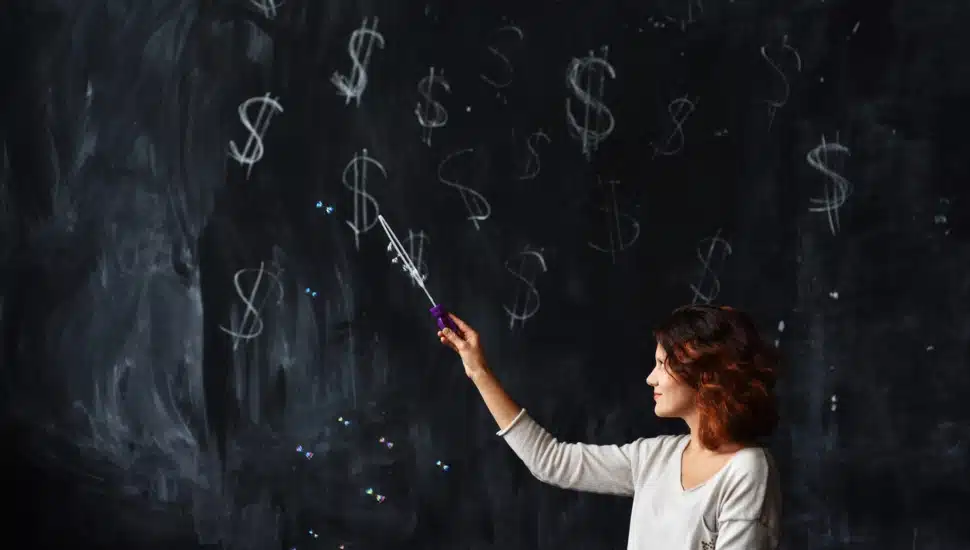 A teacher uses a pointer to point to dollar signs on a blackboard.
