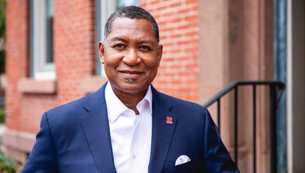 Antonio D. Tillis, Ph.D., Chancellor of Rutgers University–Camden, smiling in a navy blue suit with a Rutgers pin, standing outside a red brick building.