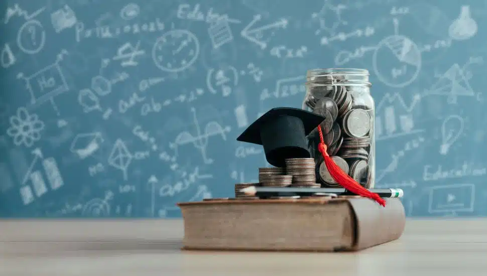 Book in front of chalkboard with a mini graduation cap and jar of money.