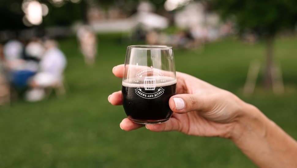 Person holding small glass of beer with Brews & Bites event logo on it.