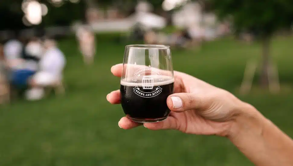 Person holding small glass of beer with Brews & Bites event logo on it.