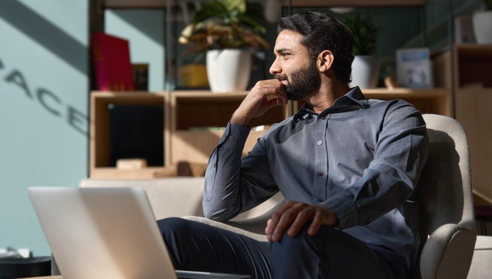 Young thoughtful smart professional business man executive looking away relaxing sitting on chair in modern office lobby with laptop.