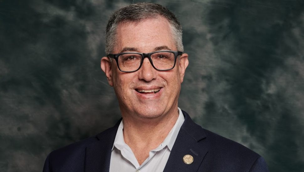 Portrait of a man wearing glasses, a navy blazer with a lapel pin, and a light-colored shirt, smiling in front of a dark, textured background.