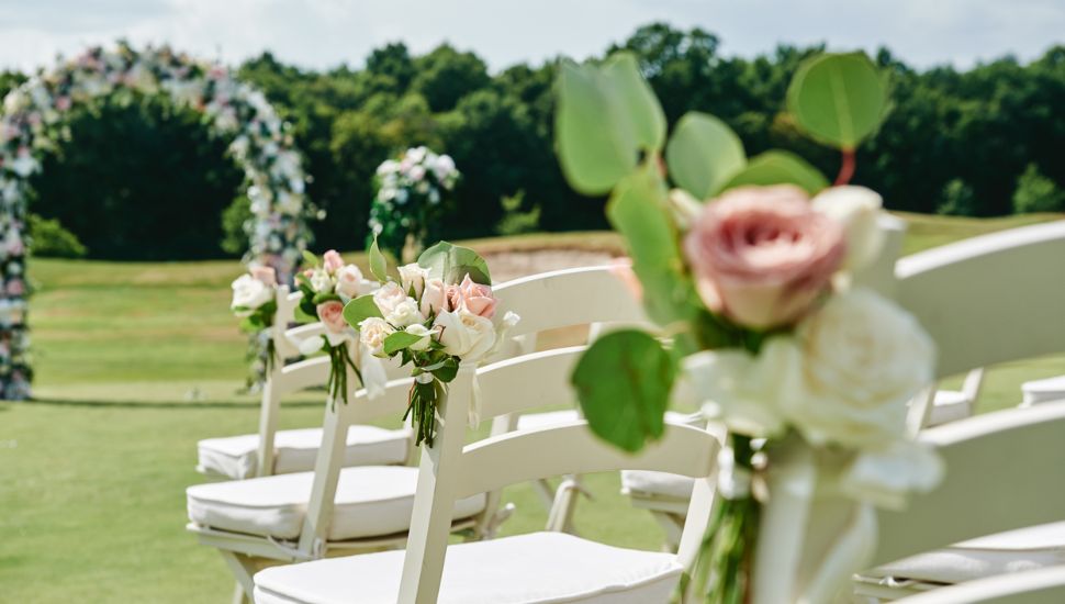 White wooden chairs with rose flowers on each side of wedding archway outdoors.