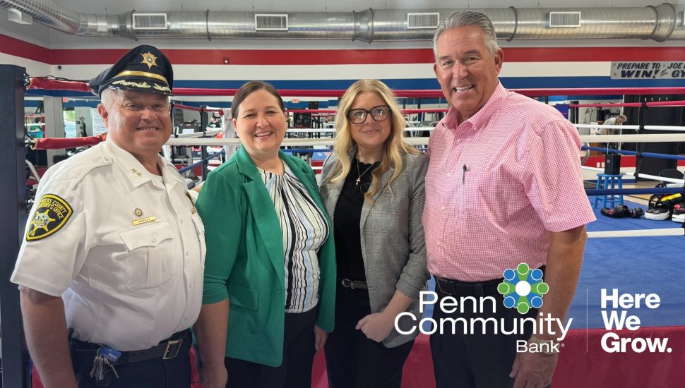 Bucks County Sheriff Frederick Harran, Penn Community Bank representatives, and Joe Hand Jr. stand together inside the Joe Hand Boxing Gym to celebrate the launch of a new youth boxing program, with a boxing ring and equipment in the background. Penn Community Bank and 'Here We Grow' logos appear in the corner.