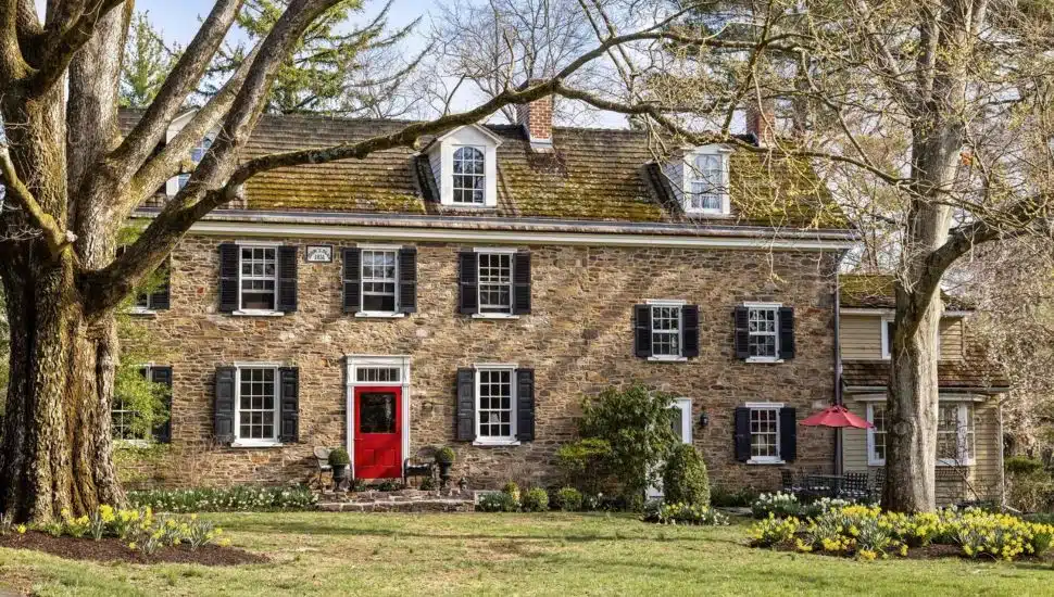 Stone manor with a red front door and a manicured lawn with old trees.