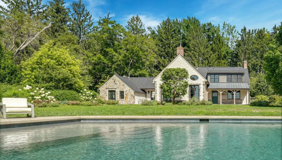 Large pool in front of a stone home partially covered by a small tree.