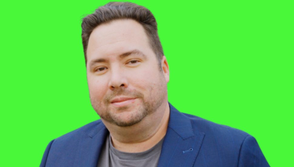 A man with short dark hair and a trimmed beard, wearing a navy blue blazer over a gray shirt, standing against a bright green background.