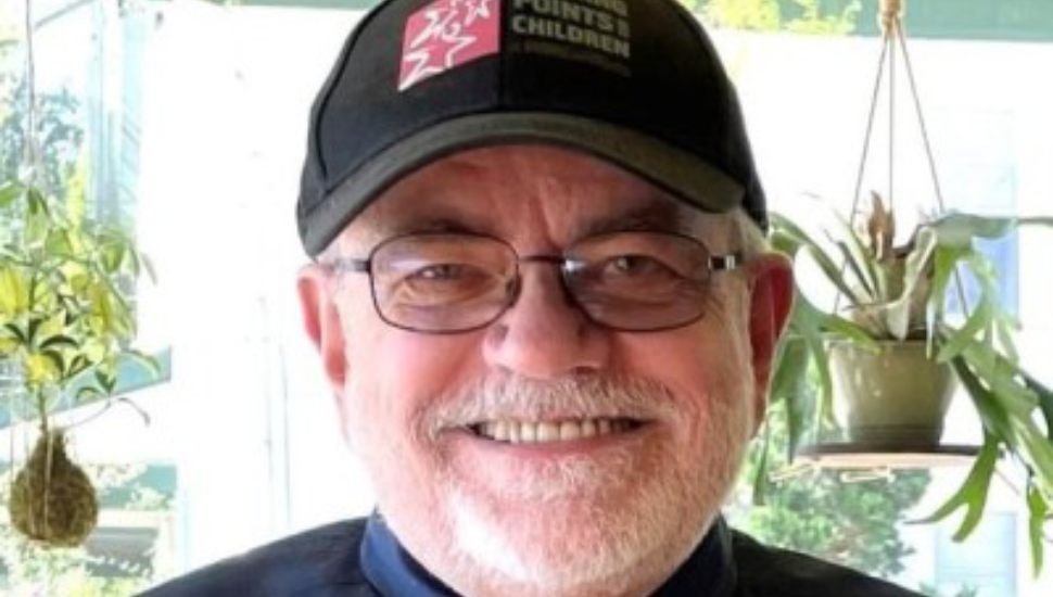 Smiling older man with glasses, a gray beard, and a black "Turning Points for Children" cap, standing indoors with hanging plants in the background.