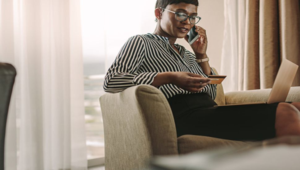 Businesswoman sitting in a chair, holding a credit card and speaking on the phone while working on a laptop.