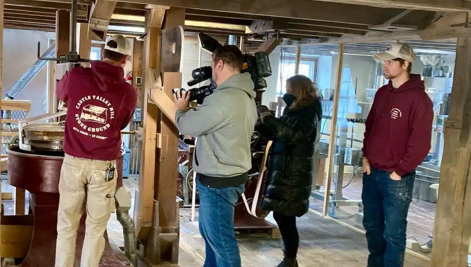 Inside a rustic wooden mill, a man in a maroon hoodie labeled 'Castle Valley Mill Stone Ground' operates milling equipment while being filmed by a cameraman. Another person holds a microphone, and a second man in a similar maroon hoodie stands nearby observing. Milling machinery and wooden beams are visible in the background.