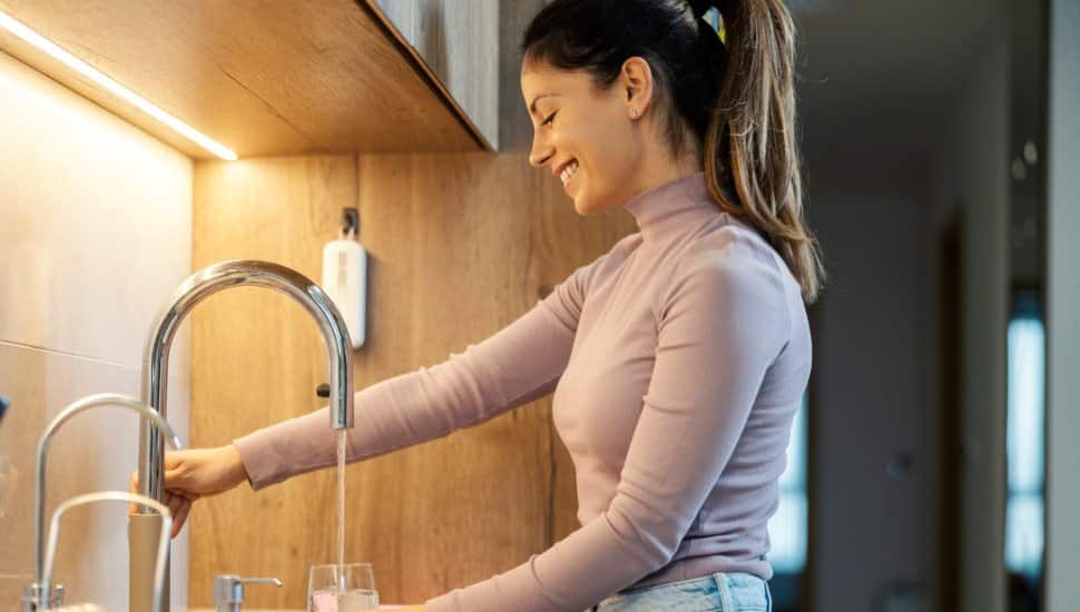 Smiling woman standing in kitchen at home and pouring clear fresh water into glass.